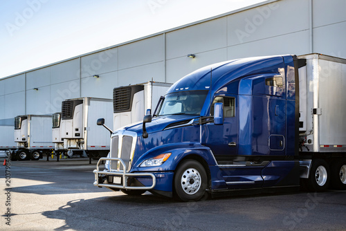 Dark blue big rig semi truck with grille guard loading cargo into refrigerated semi trailer standing in the warehouse dock gate