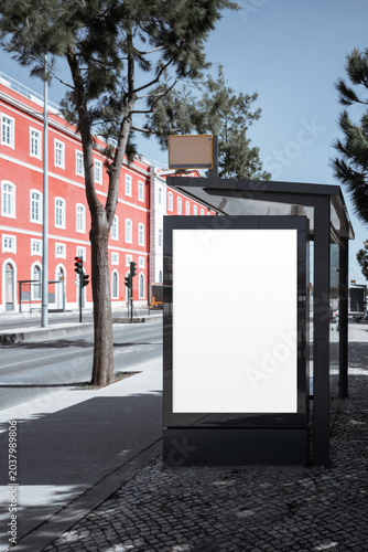 Empty backlit poster panel on black bus stop shelter beside red historic building and trees, ideal for urban advertising mockup, campaign preview or branding template