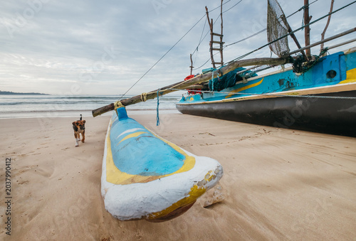 wide angle photo of traditional vintage Sri Lankan fishing boat with outrigger on sandy beach and small dog walking near sea representing local maritime heritage coastal culture and tropical island tr