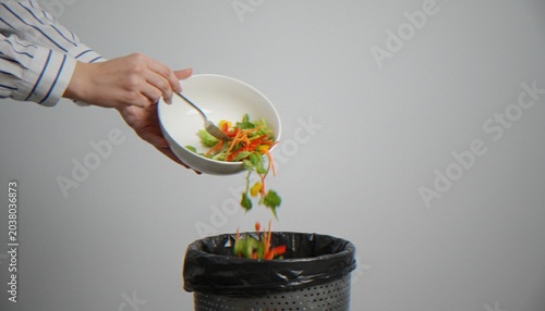 Foto A close-up shot of a person emptying a white bowl of fresh vegetable salad into a black trash bin, representing food waste and poor diet choices