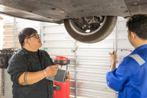Mechanic checks undercarriage with focused care. Technician smiles while holding diagnostic tool. Partner observes attentively, leaning slightly forward. Workshop tools and equipment surround them
