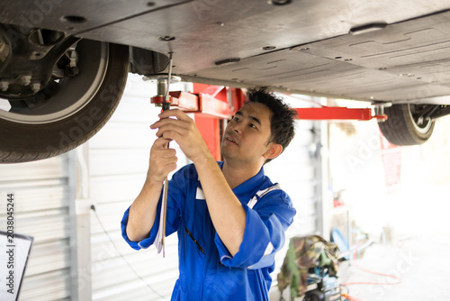 Mechanic checks undercarriage with focused care. Technician smiles while holding diagnostic tool. Partner observes attentively, leaning slightly forward. Workshop tools and equipment surround them
