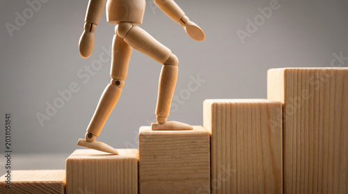 A wooden mannequin steps up a staircase made of wooden blocks on a gray background