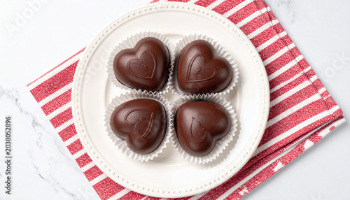 Heart-shaped Chocolates on a Plate: Four exquisite dark chocolate confections, shaped like hearts and nestled in white wrappers, are presented on a white plate with a red and white striped napkin.