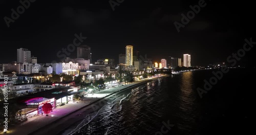 Forward aerial night view of Sihanoukville shows a vibrant coastline, where illuminated high-rise hotels and beachside businesses line the sandy shore as waves reflect the glowing cityscape.