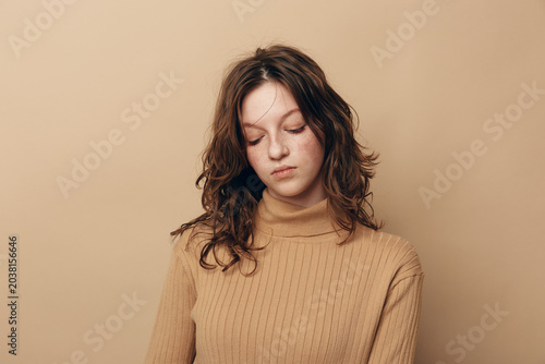 Fotografie Sad young woman with curly hair wearing beige sweater looks down with closed eyes