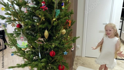 Cheerful child wearing white dress standing near decorated christmas tree, experiencing festive holiday magic with genuine happiness and sparkling wonder