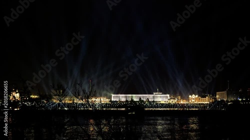 skyline illuminated with dynamic beams, lighting artists craft immersive scenes across bustling waterfront area, technical team synchronizes luminous sequences to create captivating nightscape display