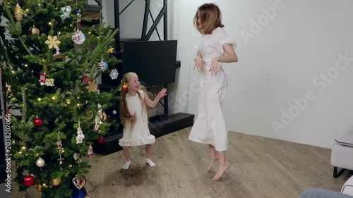 Festive white clad mother and daughter dancing near sparkling christmas tree, sharing warmth and holiday spirit in cozy living room