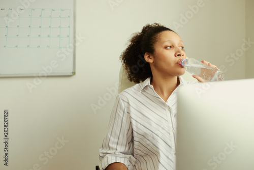 Young adult African American businesswoman drinking water while working at desk  supporting workplace wellness and hydration habits. Useful for corporate health and productivity campaigns