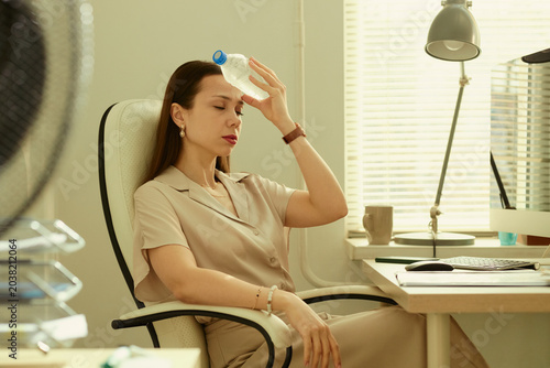 Young adult Caucasian businesswoman cooling forehead with water bottle while sitting at office desk  experiencing heat stress and fatigue. Useful for workplace wellness or summer productivity