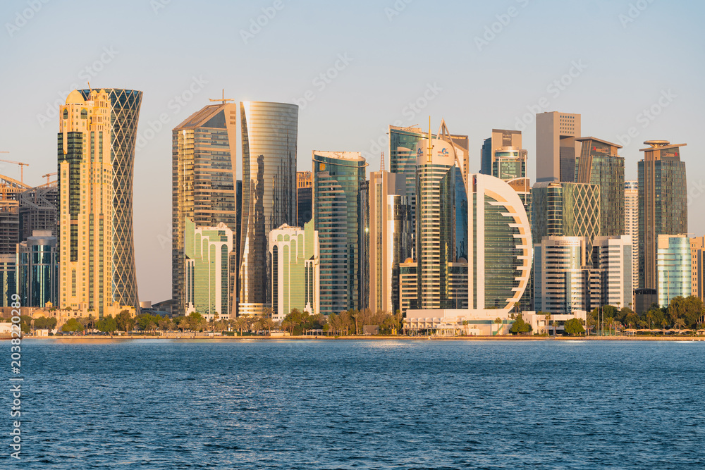 Obraz premium The modern Doha skyline at sunset with waterfront trees and blue sea.