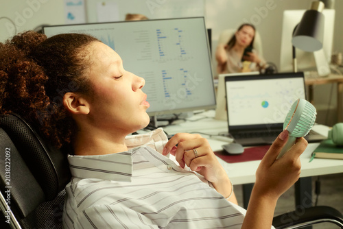 Foto Young adult woman cooling herself with handheld fan while sitting at office desk, struggling with workplace heat during computer work