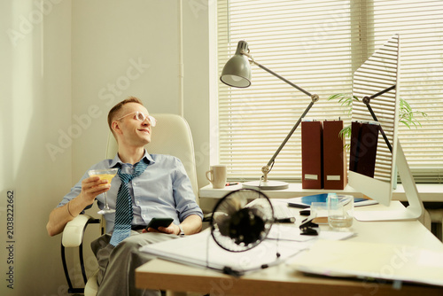 Konstfotografi Caucasian young adult businessman relaxing at office desk, holding cocktail glass and smartphone, loosening tie after work
