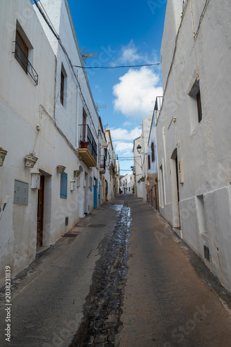 A narrow, uphill street in Nijar, Almeria, Spain, with whitewashed buildings, central cobblestone paving, flowerpots, and small balconies