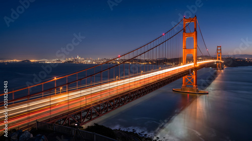 Iconic orange suspension bridge at night featuring long exposure traffic light trails with a glowing city skyline in the background.