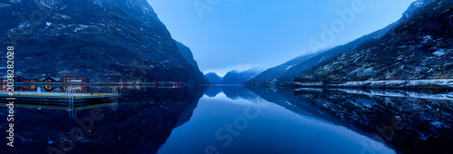 Tranquil blue hour landscape at the Norwegian Fjord in Flam, Norway, featuring perfect mirror reflections of majestic mountains in still water at early morning dawn.