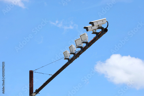 Security cameras and floodlights installed on a high pole under a bright blue sky, representing surveillance technology, public safety, monitoring systems, and urban security infrastructure.