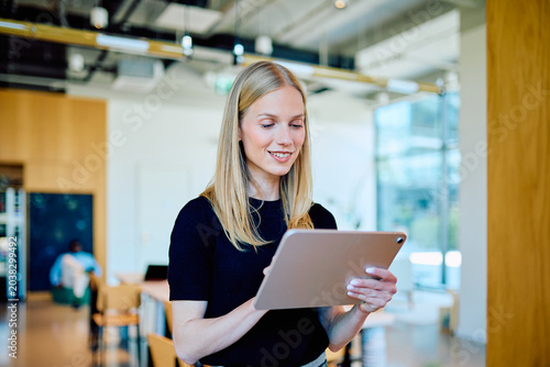 Bild auf Leinwand Young woman smiling, standing in a coworking space and working or browsing on a digital tablet