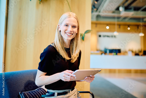 Tableau sur toile Young blonde woman smiling, working with a tablet in a modern office, representi