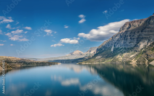Turquoise Saint Mary Lake surrounded by steep forested mountain slopes and weathered tree stumps under a bright blue Montana sky in Glacier National Park
