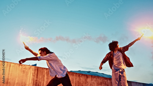 Quadro su tela In the golden glow of the setting sun, two young skateboarders, their faces alight with the thrill of the moment, ignite flares, casting vibrant trails of light against a clear blue sky