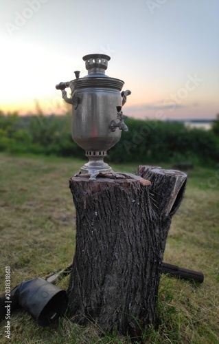 Photos Vintage metal samovar on a wooden stump outdoors at sunset