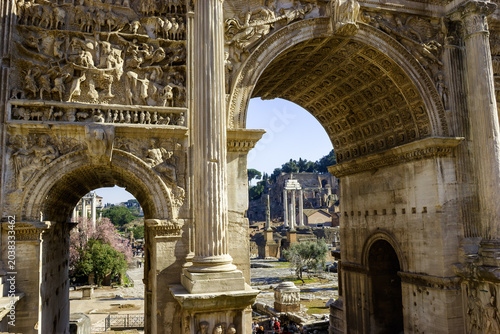 Foto The Arch of Titus in Rome, Italy, showcases intricate Roman carvings and ancient ruins, with tourists exploring the historical site under a clear blue sky