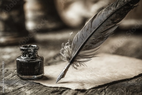 A quill pen rests atop aged paper with inkwell on a textured, wooden surface. Soft focus background
