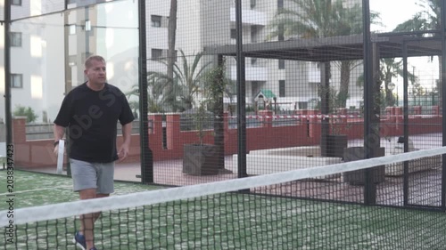 Man playing padel on an outdoor court in a residential area, raising his racket to hit the ball near the net. Casual sports lifestyle scene showing an amateur player during a padel training session or