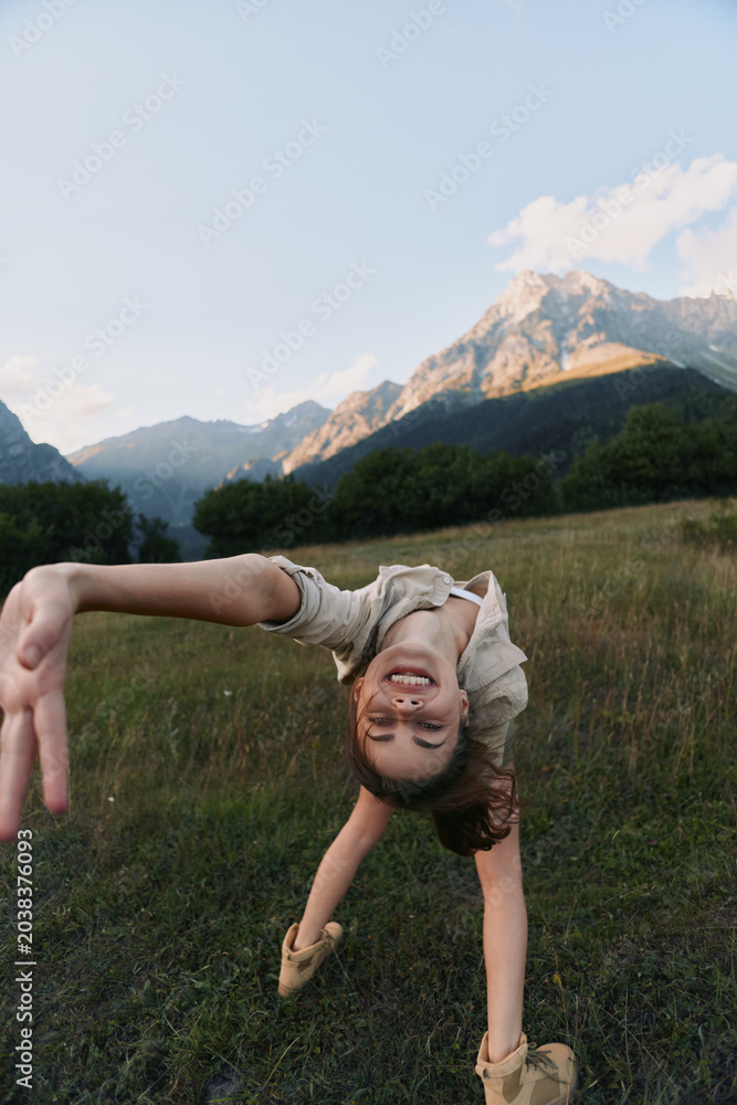 Obraz premium A smiling child performs a backbend in a meadow with mountains in the background, capturing carefree outdoor play, movement and balance in a bright nature scene.