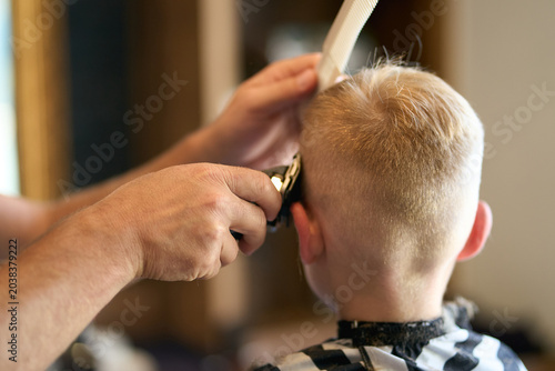 Fotografía Coiffeur coupant les cheveux d'un petit garçon blond avec une tondeuse électrique et un peigne dans un salon de coiffure ou à la maison