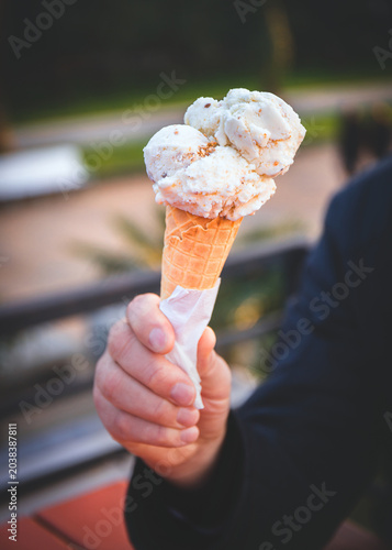 Male hand holding vanilla ice cream cone outdoors on a summer day. Sweet frozen dessert in waffle cone with creamy scoops with blurred background. Concept of street food, leisure. 