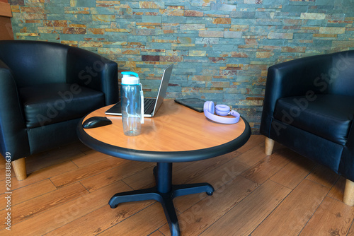 Modern workspace with laptop and headphones on round wooden table. Cozy business interior with armchairs and indoor lighting. Remote work and relaxation concept. Close-up, selective focus.