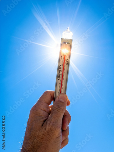 POV hand holding red thermometer up to blazing summer sun and blue sky extreme heat alert.