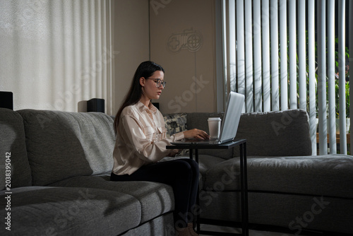 Young woman working on laptop from home daytime sitting on sofa with coffee cup nearby creating cozy remote work atmosphere and comfortable home office lifestyle