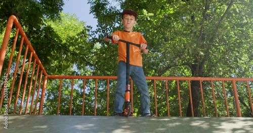 Boy twists scooter handlebar while standing on skate park platform. Child plants feet apart turning steering column to practice ride control before moving