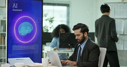 Mixed race Businessman, programmer working on a computer while looking at AI assistant interface on monitor at office. Smart workplace automation, future software engineering, machine learning concept