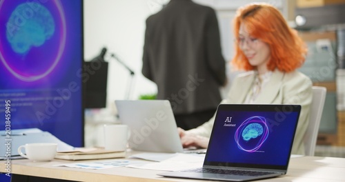 Businesswoman or programmer working on a computer while looking at AI assistant interface on monitor at office. Smart workplace automation, future software engineering, machine learning concept.