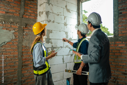 Professional construction workers and site manager inspecting brick wall structure while measuring interior building quality and checking renovation progress at construction site.