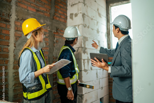 Architect and construction workers inspecting unfinished interior wall structure while reviewing safety standards and engineering measurements during onsite project audit.