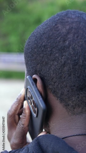 Vertical: Back view of a young African man talking on his smartphone while standing in a park during a calm and everyday outdoor moment.