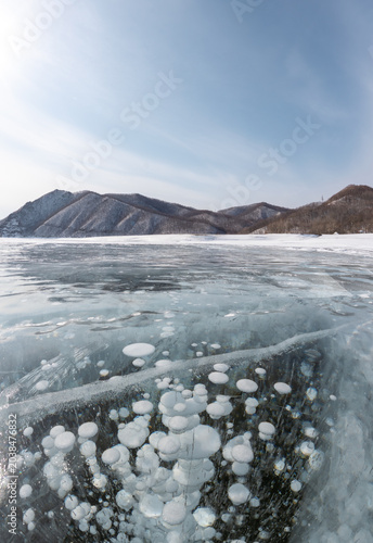 Canvas Print Ice-covered lake with frozen bubbles beneath the surface, surrounded by snow-cap