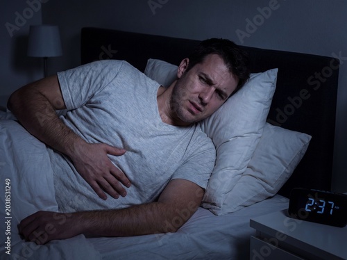 A medium shot of a man in bed late at night suffering from gastroesophageal reflux, propped up by three or four pillows to remain almost sitting with a hand resting on the upper part of his stomach, p