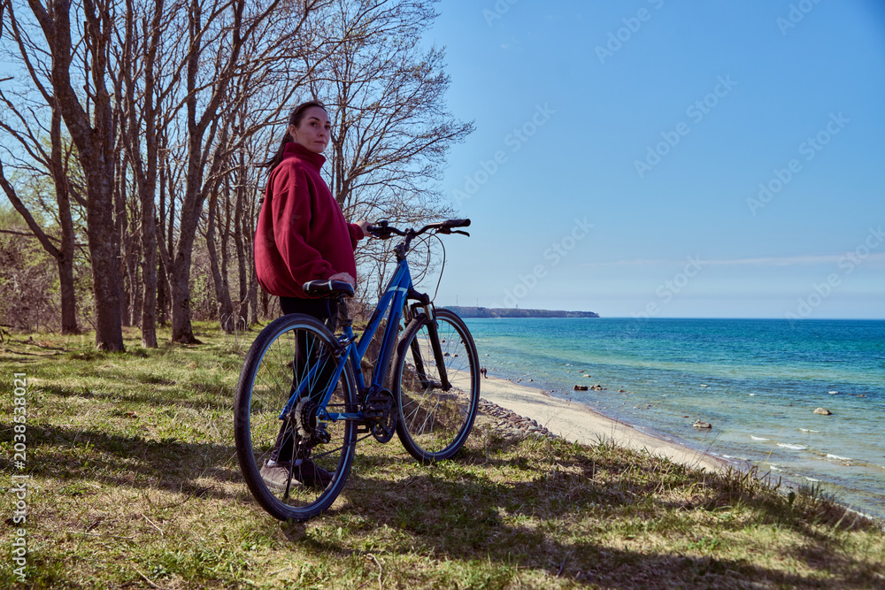 Fototapeta premium Young woman standing with bicycle near seaside under clear blue sky in sunny outdoor setting