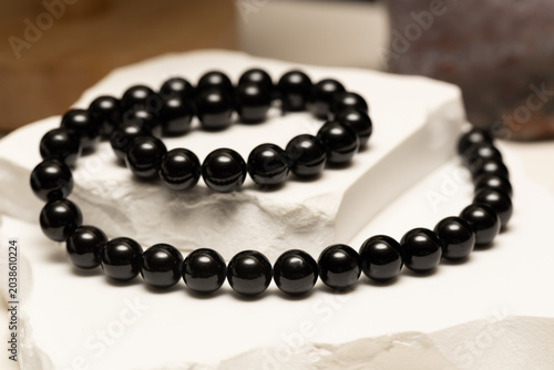 Black tourmaline beads lay on a white stone surface. The beads can be used in various jewelry items like bracelets and necklaces. They have a smooth finish and are all black.