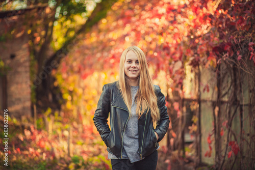 Happy blonde woman in leather jacket posing outdoors in colorful autumn park