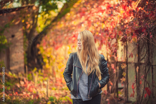 Pensive blonde woman in leather jacket against vibrant autumn foliage background
