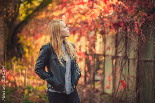 Pensive blonde woman in black jacket outdoors with vibrant autumn foliage