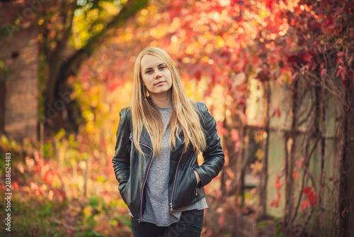 Confident young blonde woman in leather jacket posing in autumn park with colorful foliage